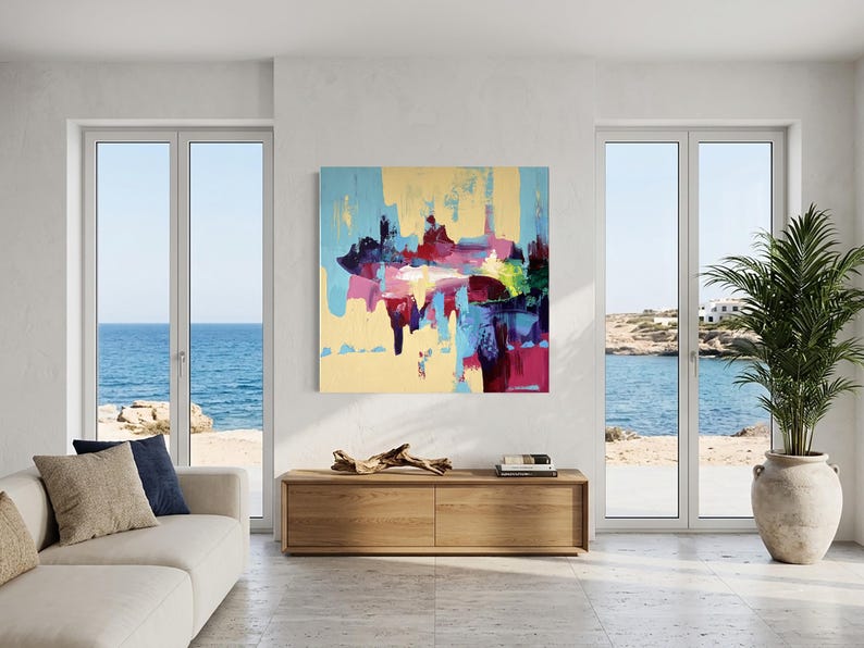 May include: An abstract painting with a light yellow background and colorful brushstrokes of blue, pink, red, and green. The artwork hangs above a wooden cabinet in a bright, airy room with a view of the ocean.