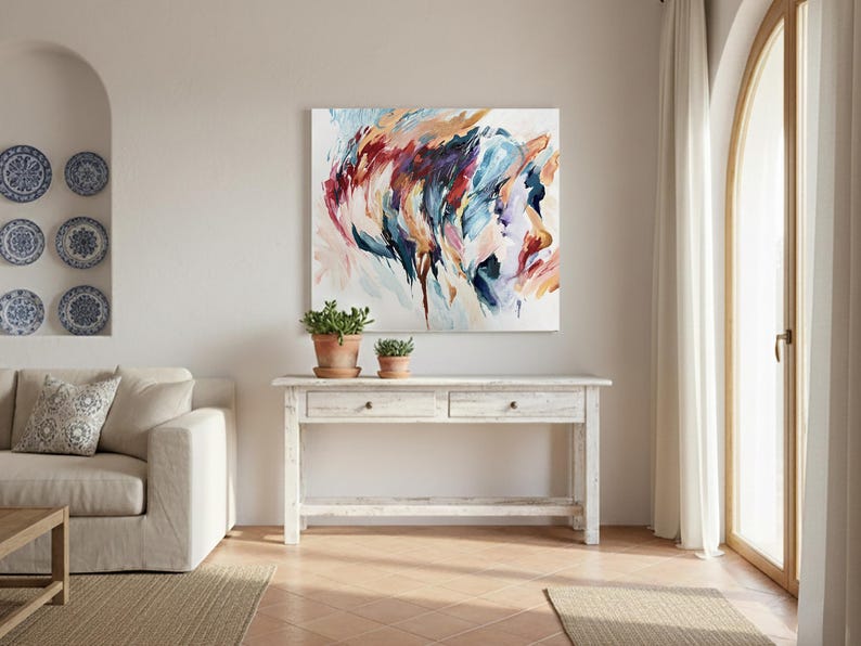 May include: Abstract art piece with a profile in shades of blue, red, and orange. The artwork hangs above a white wooden console table with two drawers. Two potted plants sit on the table. The room has a neutral color palette.