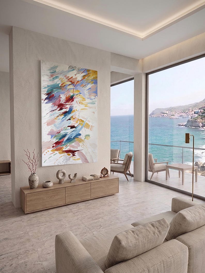 Puede incluir: Una pintura abstracta con fondo blanco y pinceladas coloridas en tonos azules, rojos, amarillos y rosas. La obra cuelga sobre un mueble de madera con objetos decorativos. La habitaci&oacute;n ofrece vistas al mar.