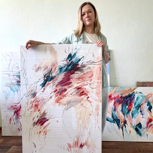 May include: Abstract art canvas with a white background and colorful brushstrokes in shades of red, teal, and beige. The artwork is held by a person, with other similar paintings visible in the background. The art is displayed on a wooden floor.