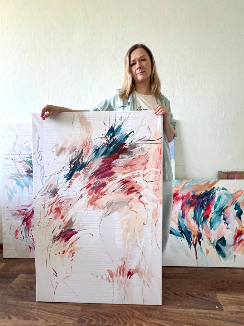 May include: Abstract art canvas with a white background and colorful brushstrokes in shades of red, teal, and beige. The artwork is held by a person, with other similar paintings visible in the background. The art is displayed on a wooden floor.