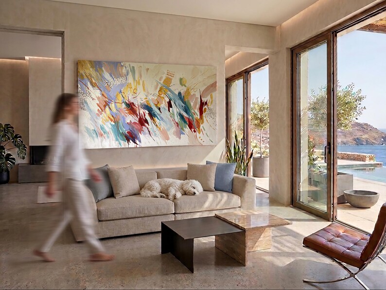 Puede incluir: Sal&oacute;n moderno con una gran pintura abstracta sobre un sof&aacute; beige. Un perro blanco descansa en el sof&aacute;. Dos mesas de centro cuadradas y una silla de cuero marr&oacute;n est&aacute;n frente a una gran ventana con vista al mar.