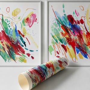May include: Two framed rectangular wall art prints featuring abstract watercolor brushstrokes in vibrant red, blue, green, and yellow hues. A rolled paper print with matching colorful abstract patterns is positioned in the foreground.