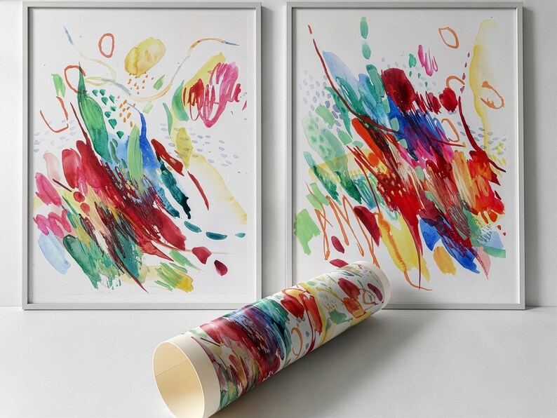May include: Two framed rectangular wall art prints featuring abstract watercolor brushstrokes in vibrant red, blue, green, and yellow hues. A rolled paper print with matching colorful abstract patterns is positioned in the foreground.