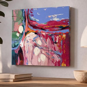 May include: Square abstract painting with a textured surface. The artwork features a blend of colors, including pink, red, blue, and green. White circular shapes are scattered across the canvas. The painting is mounted on a wall above a stack of books and a decorative bowl.