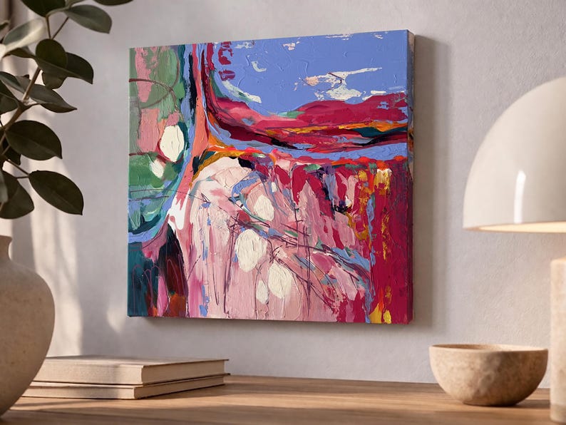May include: Square abstract painting with a textured surface. The artwork features a blend of colors, including pink, red, blue, and green. White circular shapes are scattered across the canvas. The painting is mounted on a wall above a stack of books and a decorative bowl.