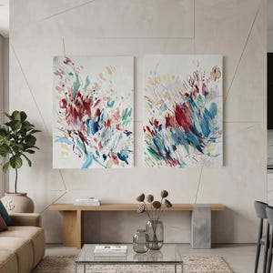 Op de afbeelding: Twee rechthoekige wandkunstdoeken met abstracte penseelstreken in tinten dieprood, blauwgroen, blauw en zachtgeel op een witte achtergrond. De schilderijen hangen naast elkaar aan een lichte muur boven een houten bank.