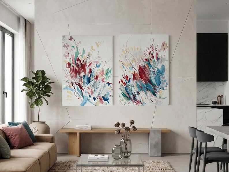 Op de afbeelding: Twee rechthoekige wandkunstdoeken met abstracte penseelstreken in tinten dieprood, blauwgroen, blauw en zachtgeel op een witte achtergrond. De schilderijen hangen naast elkaar aan een lichte muur boven een houten bank.