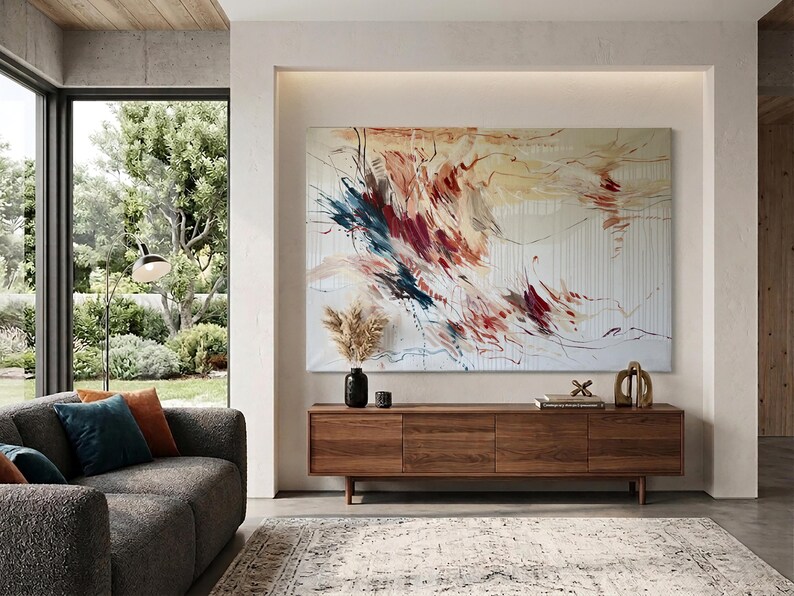 May include: A modern living room featuring a large abstract painting with red, blue, and yellow brushstrokes. Below the artwork is a wooden cabinet with three doors, and a gray sofa with colorful throw pillows. A patterned rug and large windows complete the scene.