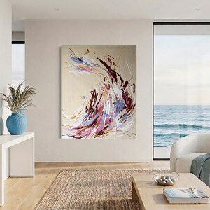 May include: Abstract art painting with a beige background and dynamic brushstrokes in shades of purple, burgundy, white, and gold. The artwork is displayed in a modern living room with a view of the ocean. A blue vase with dried plants sits on a white table.