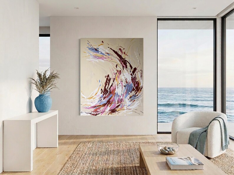 May include: Abstract art painting with a beige background and dynamic brushstrokes in shades of purple, burgundy, white, and gold. The artwork is displayed in a modern living room with a view of the ocean. A blue vase with dried plants sits on a white table.