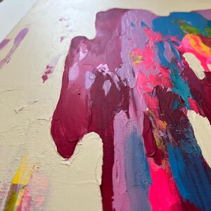 May include: Abstract artwork with bold brushstrokes in pink, magenta, blue, and yellow on a cream backdrop. The painting has a textured surface, with visible paint layers and a dynamic composition. The style is modern and vibrant, with a focus on colour and texture.