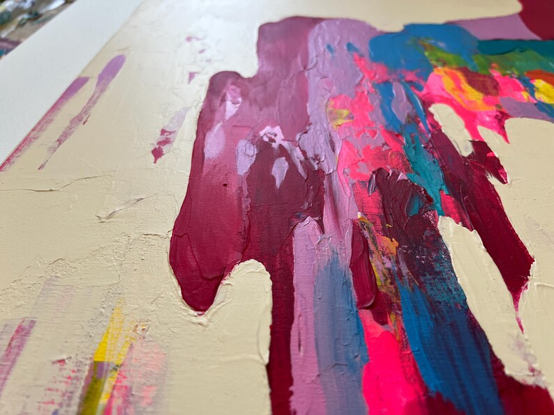May include: Abstract artwork with bold brushstrokes in pink, magenta, blue, and yellow on a cream backdrop. The painting has a textured surface, with visible paint layers and a dynamic composition. The style is modern and vibrant, with a focus on colour and texture.