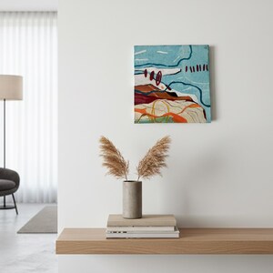 May include: Abstract landscape painting with a textured surface. The artwork features a combination of colors, including blue, red, orange, and white. The painting is mounted on a white wall above a wooden shelf. A vase with dried plants sits on the shelf.