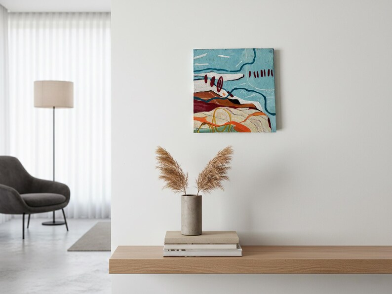 May include: Abstract landscape painting with a textured surface. The artwork features a combination of colors, including blue, red, orange, and white. The painting is mounted on a white wall above a wooden shelf. A vase with dried plants sits on the shelf.