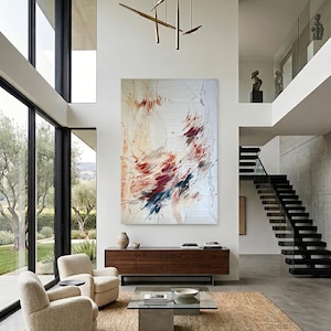 May include: Modern living room with large windows, a neutral color palette, and an abstract painting. Two cream-colored armchairs face a glass coffee table and a wooden cabinet. A woven rug covers the floor.