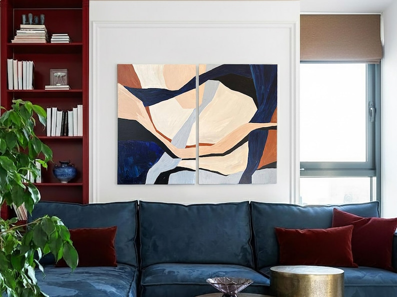 May include: Abstract art diptych with a modern design. The artwork features a combination of shapes and colors, including navy blue, beige, and rust. The painting is displayed above a blue sectional sofa with burgundy throw pillows.