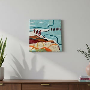 May include: An abstract painting with a textured surface. The artwork features a landscape-like composition with shades of blue, brown, and orange. White and yellow lines add detail. The painting is displayed on a white wall above a wooden cabinet.
