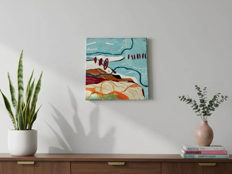 May include: An abstract painting with a textured surface. The artwork features a landscape-like composition with shades of blue, brown, and orange. White and yellow lines add detail. The painting is displayed on a white wall above a wooden cabinet.