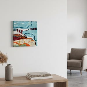 May include: Abstract art piece with a textured surface. The artwork features a light blue background with red, orange, and white shapes. The piece is mounted on a square canvas and hangs on a white wall. A vase with dried plants and books are on a wooden table.