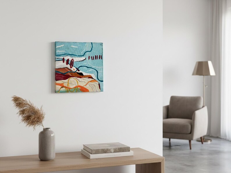 May include: Abstract art piece with a textured surface. The artwork features a light blue background with red, orange, and white shapes. The piece is mounted on a square canvas and hangs on a white wall. A vase with dried plants and books are on a wooden table.