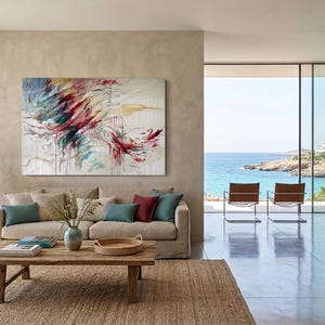May include: An abstract painting with red, blue, and yellow brushstrokes hangs above a beige sofa with teal and burgundy pillows. A wooden coffee table sits on a woven rug. The room has a large window overlooking the ocean.