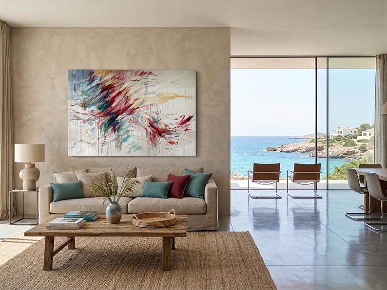 May include: An abstract painting with red, blue, and yellow brushstrokes hangs above a beige sofa with teal and burgundy pillows. A wooden coffee table sits on a woven rug. The room has a large window overlooking the ocean.
