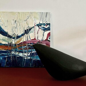 May include: An abstract painting with thick brushstrokes in white, blue, pink, and orange hues. A black, bird-shaped sculpture sits in front of the painting on a red surface. The artwork is displayed against a white wall.
