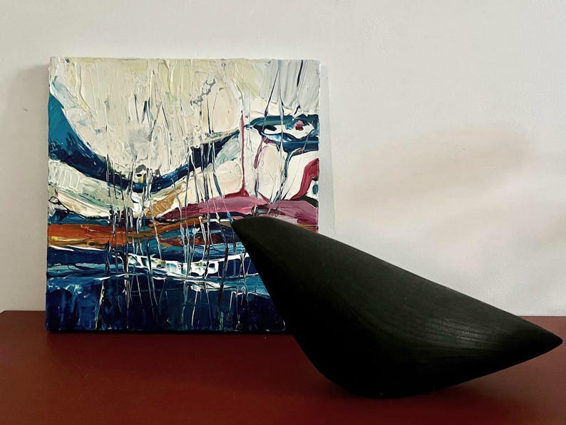 May include: An abstract painting with thick brushstrokes in white, blue, pink, and orange hues. A black, bird-shaped sculpture sits in front of the painting on a red surface. The artwork is displayed against a white wall.