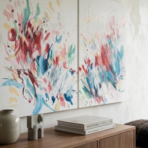 Op de afbeelding: Twee rechthoekige doeken met abstracte penseelstreken in tinten rood, blauwgroen, roze en geel op een witte achtergrond. Het kunstwerk hangt boven een houten consoletafel met een keramische vaas, een klein beeldhouwwerk en twee boeken.