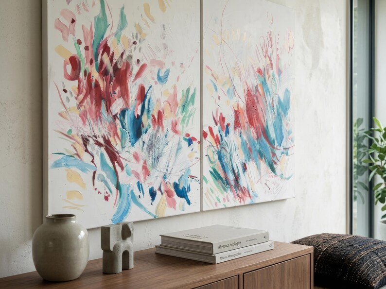 Op de afbeelding: Twee rechthoekige doeken met abstracte penseelstreken in tinten rood, blauwgroen, roze en geel op een witte achtergrond. Het kunstwerk hangt boven een houten consoletafel met een keramische vaas, een klein beeldhouwwerk en twee boeken.