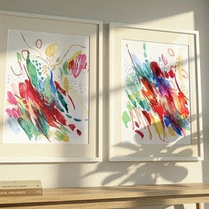 May include: Two framed wall art prints featuring abstract watercolor paintings with vibrant splashes of red, blue, green, and yellow brushstrokes on a white background. The prints are housed in matching light-colored frames with white matting.