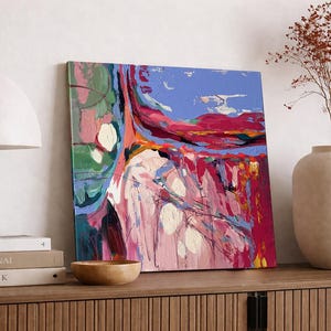 May include: Abstract art painting with a vibrant mix of colors, including pink, blue, red, and green. The artwork is displayed on a wooden surface, alongside a lamp, books, a wooden bowl, and a vase with dried flowers. The painting has a textured appearance.