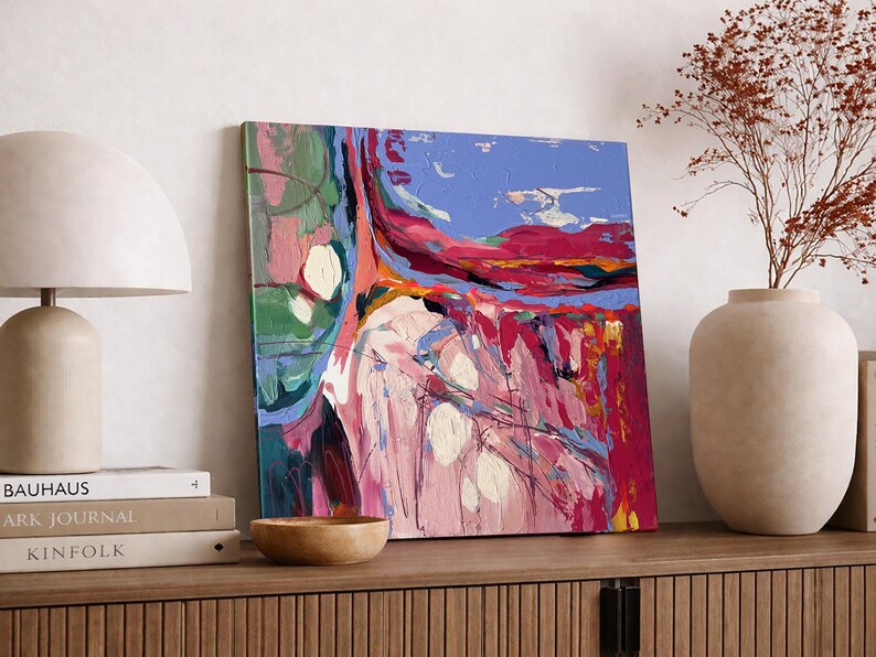 May include: Abstract art painting with a vibrant mix of colors, including pink, blue, red, and green. The artwork is displayed on a wooden surface, alongside a lamp, books, a wooden bowl, and a vase with dried flowers. The painting has a textured appearance.