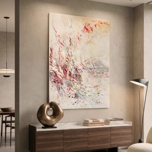 May include: A large vertical abstract painting with red, pink, and green brushstrokes on a cream background, displayed above a dark wood sideboard with a bronze sculptural object and two books, one titled Rothko, resting on the white marble top.