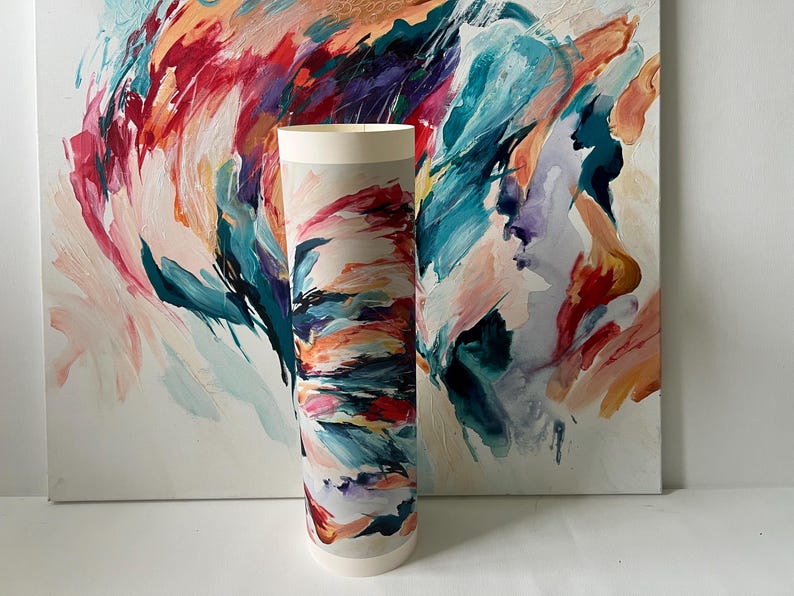 K&ouml;nnte beinhalten: Ein zylindrischer Papierdruck mit einem lebendigen abstrakten Gem&auml;lde, das kr&auml;ftige, ausdrucksstarke Pinselstriche in T&uuml;rkis, Dunkelblau, Magenta, Orange und Creme zeigt. Der Druck steht aufrecht vor einer passenden gro&szlig;en quadratischen Leinwand.