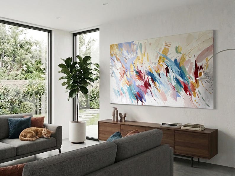 Puede incluir: Un sal&oacute;n moderno con una gran pintura abstracta con vibrantes pinceladas en rojo, azul, amarillo y blanco. Un mueble de madera oscura alberga objetos decorativos y libros. Un sof&aacute; gris con cojines azules y naranjas y un gato naranja durmiendo.