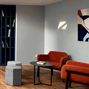 May include: Interior shot of a modern living space with a blue bookshelf, two orange armchairs, and a small black table. Two gray ottomans sit in front of the chairs. A painting with abstract shapes hangs on the wall.