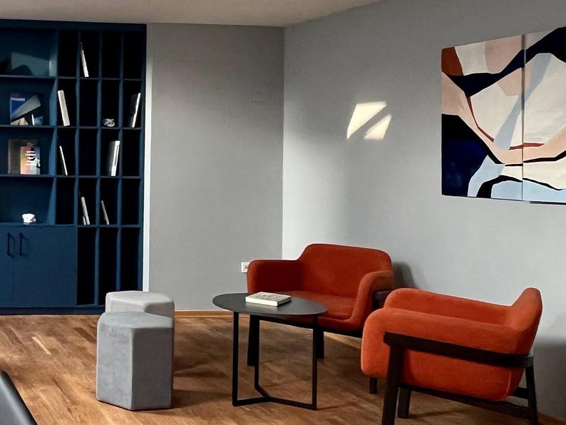 May include: Interior shot of a modern living space with a blue bookshelf, two orange armchairs, and a small black table. Two gray ottomans sit in front of the chairs. A painting with abstract shapes hangs on the wall.