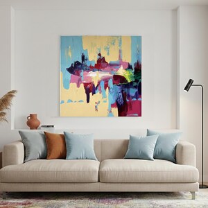 May include: An abstract painting with a yellow background and splashes of blue, red, and purple hangs above a beige sofa. The sofa has three light blue and one brown pillow. A wooden side table holds a blue vase with dried plants.