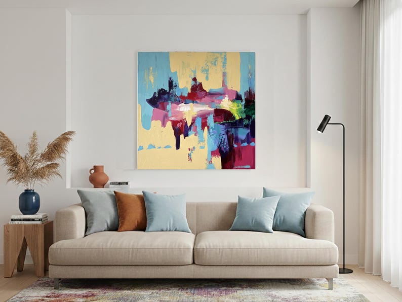May include: An abstract painting with a yellow background and splashes of blue, red, and purple hangs above a beige sofa. The sofa has three light blue and one brown pillow. A wooden side table holds a blue vase with dried plants.