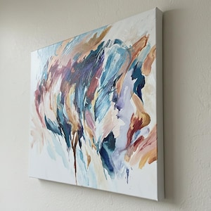 May include: Square canvas art with an abstract painting. The artwork features a blend of colors, including blues, purples, and oranges, with brushstrokes and drips. The canvas is mounted on a white frame, creating a modern aesthetic.