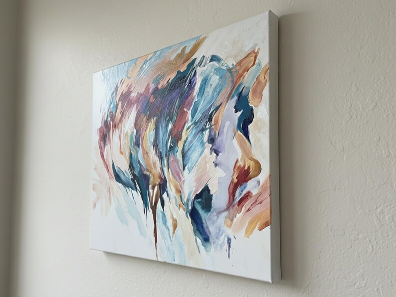 May include: Square canvas art with an abstract painting. The artwork features a blend of colors, including blues, purples, and oranges, with brushstrokes and drips. The canvas is mounted on a white frame, creating a modern aesthetic.