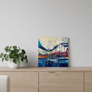 May include: Abstract oil painting on canvas with a textured surface. The artwork features a landscape with blue, white, orange, and red hues. The painting is displayed on a light wood cabinet with a potted plant and a white vase.
