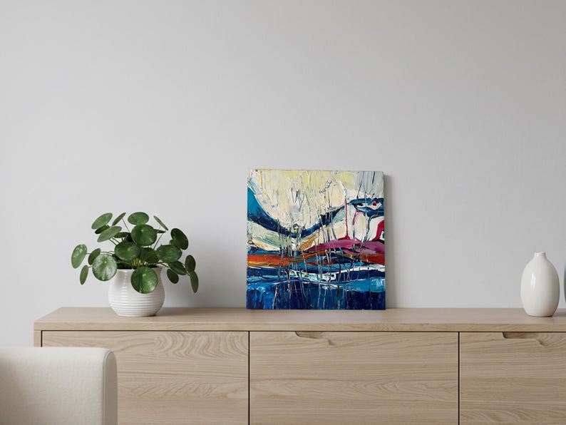 May include: Abstract oil painting on canvas with a textured surface. The artwork features a landscape with blue, white, orange, and red hues. The painting is displayed on a light wood cabinet with a potted plant and a white vase.