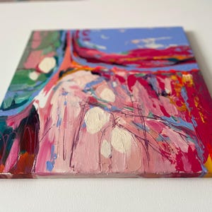 May include: Square abstract painting with thick brushstrokes in shades of pink, red, blue, green, and white. The artwork has a textured appearance, with visible paint layers and a vibrant color palette. The canvas is mounted on a wooden frame.