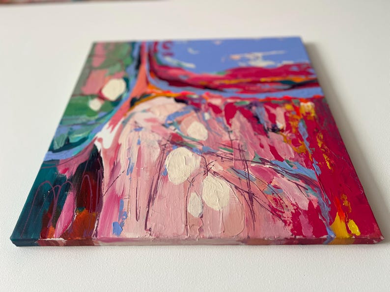 May include: Square abstract painting with thick brushstrokes in shades of pink, red, blue, green, and white. The artwork has a textured appearance, with visible paint layers and a vibrant color palette. The canvas is mounted on a wooden frame.