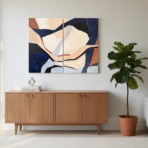 May include: Abstract art piece in a living room setting. The artwork is split into two panels, featuring a design with navy, beige, and rust tones. A wooden cabinet with four doors sits below the art. A potted plant and a sofa complete the scene.