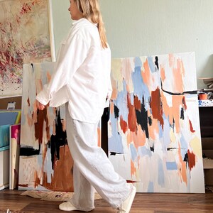 May include: A long-sleeved white button-down shirt with a collar and two chest pockets, paired with loose-fitting light grey trousers and white trainers. The outfit is worn by a person standing in front of large abstract paintings with orange, blue, black, and white brushstrokes.