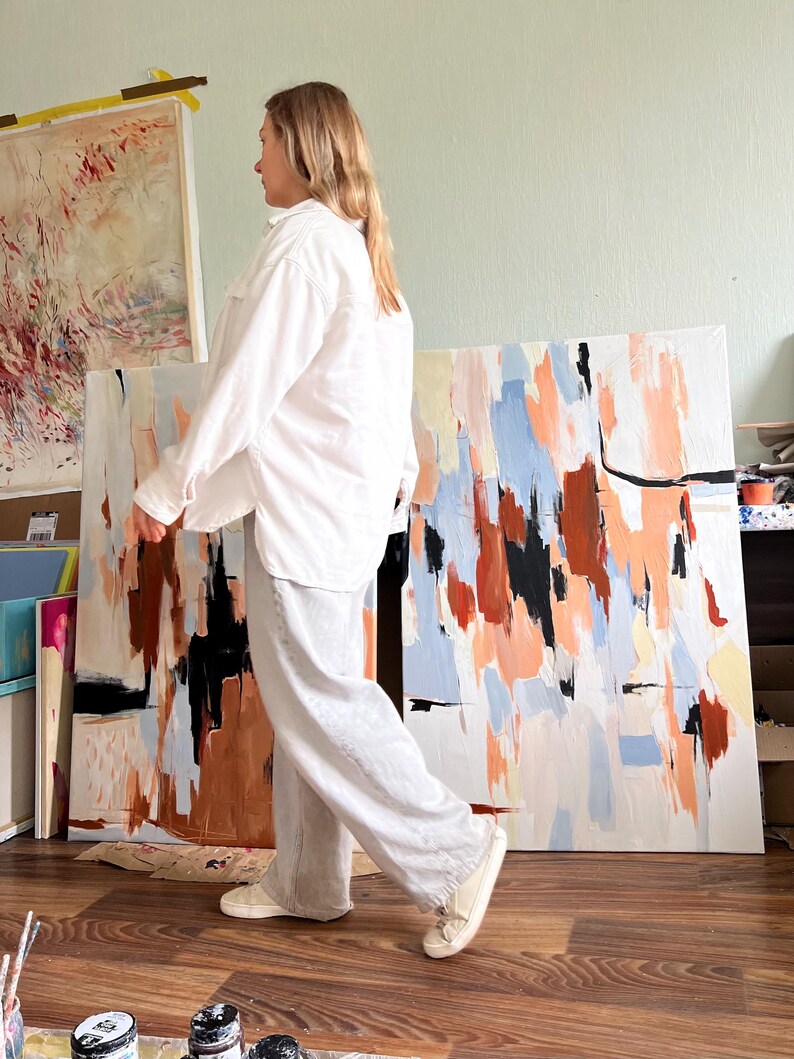 May include: A long-sleeved white button-down shirt with a collar and two chest pockets, paired with loose-fitting light grey trousers and white trainers. The outfit is worn by a person standing in front of large abstract paintings with orange, blue, black, and white brushstrokes.
